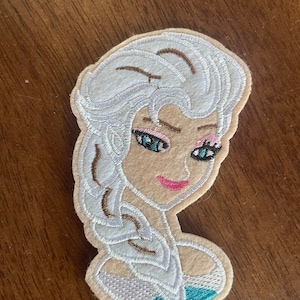 May include: White felt embroidered patch of a cartoon character with blue eyes, pink lips, and a white braid. The character is wearing a light blue and white dress.