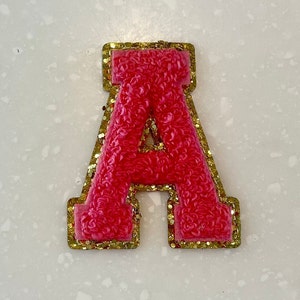 May include: A pink felt letter A with a gold glitter outline. The letter is a decorative patch or applique.