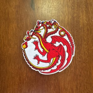 May include: Embroidered patch featuring a red three-headed dragon design with yellow accents on a white background. The patch has a detailed, textured appearance and a stitched border. Ideal for jackets, bags, or other fabric items.
