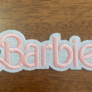 May include: Pink felt patch with the word "Barbie" embroidered in a white outline.