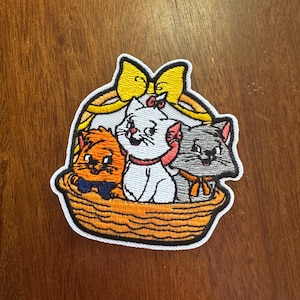 May include: Embroidered patch featuring three cartoon kittens in a woven basket. The kittens are white, orange, and gray, each with a bow. A yellow bow tops the basket. The patch has a white border.
