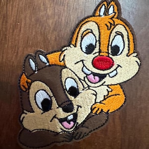 May include: Embroidered patch featuring Chip and Dale, the Disney chipmunk duo. Chip is brown, and Dale is orange, both with white and black eyes, red noses, and smiling with their tongues out. The patch has a brown border.