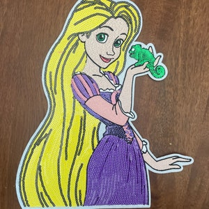 May include: A sequined patch featuring a character with long yellow hair, wearing a purple and pink dress, holding a small green chameleon. The design is outlined in white and captures the character from the waist up with a smiling expression.