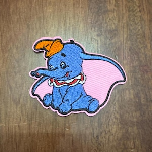 May include: Embroidered iron-on patch featuring a cartoon Dumbo the elephant wearing a pink hat and a red and white striped collar. The patch is on a pink background.