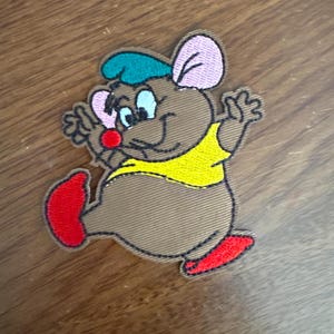May include: An embroidered patch featuring a cartoon mouse wearing a blue and green hat, a yellow shirt, and red shoes. The mouse is smiling and waving.