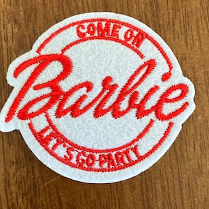 May include: A white felt patch with red embroidered text. The patch is circular with the word "Barbie" in a cursive font in the center. The words "COME ON" and "LET'S GO PARTY" are in a red font around the edge of the patch.