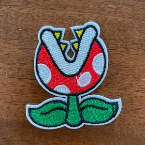 May include: A red and green embroidered patch of a plant with a large mouth, resembling a Piranha Plant from the Super Mario video game series.