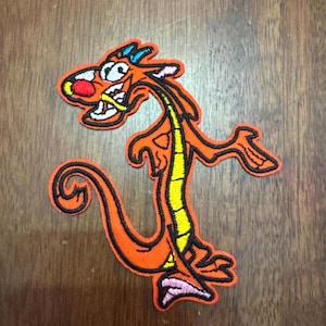 May include: Orange embroidered iron-on patch of a cartoon dragon with a yellow spine, red nose, and blue and white eyes.