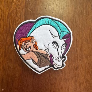May include: Embroidered patch featuring a cartoon image of a character with red hair and a stern expression, alongside a white horse-like creature. The patch is heart-shaped with a purple border and teal and white accents.