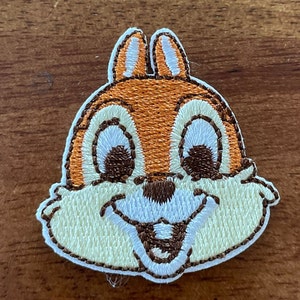 May include: Embroidered patch featuring a cartoon squirrel with brown fur, white eyes, and a big smile. The patch is white with brown stitching around the edges.