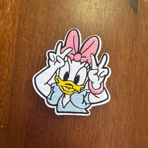 May include: Embroidered patch featuring Daisy Duck, a cartoon character, making a peace sign with both hands. Daisy Duck is wearing a pink bow, a light blue top, and a pink bracelet. The patch has a white border.