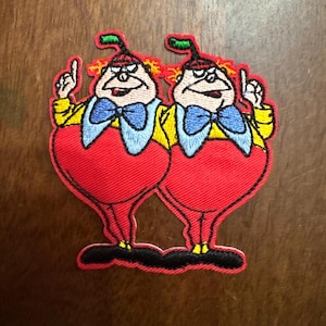 May include: Embroidered patch featuring two cartoon characters in red, yellow, and blue. The figures have red heart-shaped bodies, yellow jackets, and blue bow ties. They are pointing upwards with their index fingers.