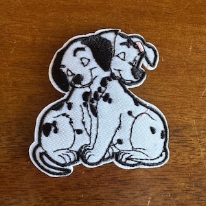 May include: Embroidered patch featuring two Dalmatian puppies. The dogs are white with black spots and black ears. The patch has a white border and is likely for application on clothing or accessories.