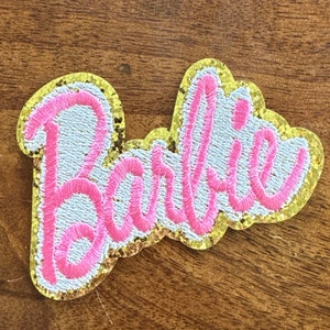 May include: Embroidered patch featuring the name "Barbie" in pink script lettering. The text is stitched onto a light blue background, outlined with gold glitter and a gold border. The patch is likely for clothing or accessories.