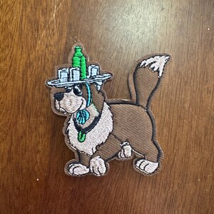 May include: Embroidered patch featuring a cartoon dog wearing a hat with a green bottle on top. The dog has brown fur with a cream-colored chest and face. The patch has a black border and a blue scarf.