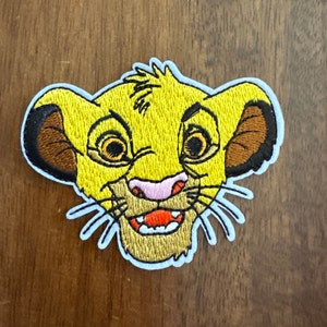 May include: A yellow embroidered patch of Simba, a lion cub from Disney's The Lion King, with a brown ear and a pink nose. Simba is smiling with his mouth open, showing his teeth.