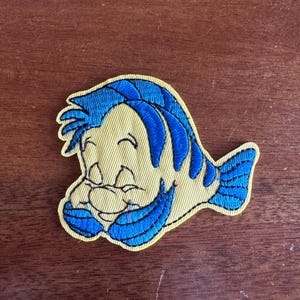 May include: Embroidered patch featuring a cartoon fish character with yellow and blue colors. The fish has blue hair and fins, and a smiling expression. The patch has a yellow border.