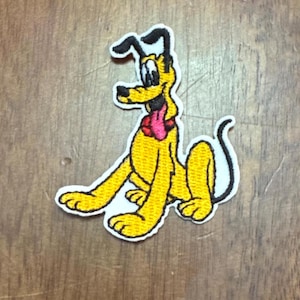 May include: An embroidered iron-on patch featuring a cartoon image of Pluto, a yellow dog with black ears and a red bow tie. Pluto is sitting with his tongue sticking out.