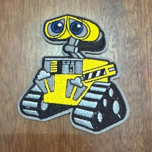 May include: A yellow and gray embroidered patch of a robot with large blue eyes and treads. The robot is from the movie Wall-E.