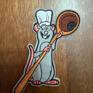 May include: Embroidered iron-on patch featuring a cartoon rat wearing a chef's hat and holding a wooden spoon. The rat is gray, the hat is white, and the spoon is brown with a black bowl.