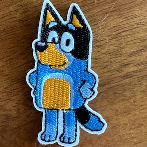 May include: Embroidered patch of a blue and black cartoon dog with a yellow chest and white eyes. The dog is standing with its arms crossed and has a friendly expression.
