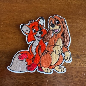 May include: Embroidered patch featuring a fox and a dog, both with smiling expressions. The fox is primarily orange with white and black accents. The dog is brown with tan and white details. The patch has a white border.