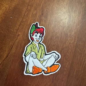 May include: Embroidered patch featuring Peter Pan, with red hair and a green hat and shirt. He is seated with orange shoes and white pants. The patch has a white border.