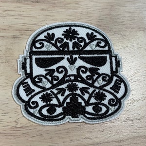 May include: Embroidered patch featuring a stylized Stormtrooper helmet design. The helmet is white with intricate black floral and ornamental detailing, and a silver border. The patch is likely for clothing or accessories.