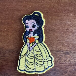 May include: Embroidered patch featuring a cartoon depiction of a princess in a yellow ball gown. The patch has black outlines and details, including the princess's black hair and red lips. The princess is holding a small orange object.