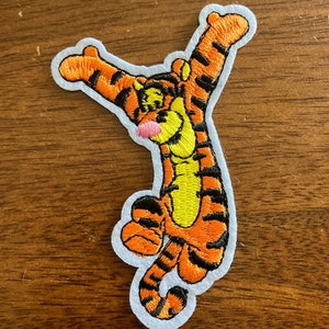 May include: An embroidered iron-on patch featuring Tigger from Winnie the Pooh. Tigger is orange and black with a yellow belly and is depicted with his arms raised in the air.