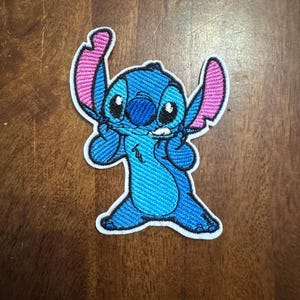 May include: Embroidered patch featuring the character Stitch from Lilo & Stitch. The patch has a blue body with pink ears and a white border. The character is in a playful pose.
