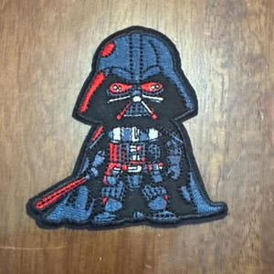May include: A black and red embroidered patch of a cartoon Darth Vader with a lightsaber. The patch is about 3 inches tall and 2 inches wide.