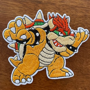 May include: Embroidered patch of Bowser, a video game character, in a yellow, orange, red, green, and black color scheme. Bowser is a large, reptilian creature with a spiky shell, a red mohawk, and a menacing expression.