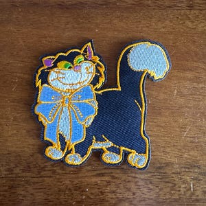 May include: A black cat patch with a blue bow tie and a smiling face. The cat has yellow, purple, and green accents. The patch is outlined in yellow and has a denim-like texture.