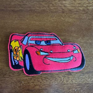 May include: Embroidered patch featuring Lightning McQueen from the Cars movie. The patch is primarily red with black, blue, yellow, and purple accents. The car has a smiling face and the number 95 on the side.