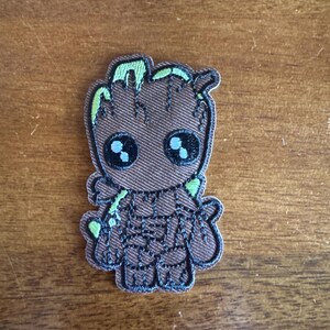 May include: A brown embroidered patch featuring a cartoon character with large black eyes and green accents. The patch has a textured appearance and is likely designed to be ironed or sewn onto fabric. The character has a tree-like appearance.