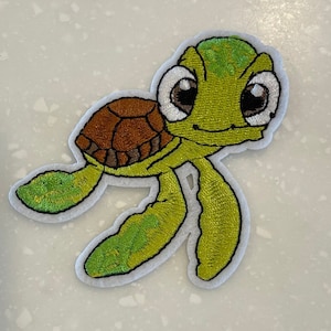 May include: Embroidered patch featuring a cartoon sea turtle. The turtle has a green body, brown shell, and large, expressive eyes. The patch is outlined in white.