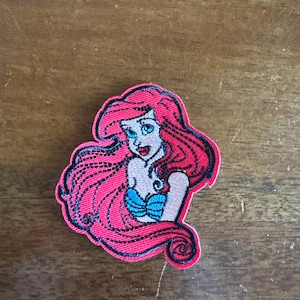 May include: Embroidered patch featuring a cartoon mermaid with long red hair, blue eyes, and a blue seashell bra. The patch has a red border and is likely for clothing or accessories.