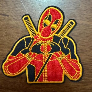 May include: Embroidered patch featuring Deadpool in a red and black suit, making a heart shape with his hands. He is holding two swords behind his back. The patch has yellow stitching.