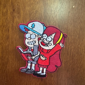 May include: Embroidered patch featuring two cartoon characters from the animated series Gravity Falls. One character wears a blue hat and the other a red dress. The patch has a red border and is on a brown surface.