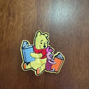 May include: Embroidered patch featuring Winnie the Pooh and Piglet reading books. Pooh is yellow with a red scarf, Piglet is pink and wearing a pink hat. The books are blue and orange with white pages. The patch has a yellow border.