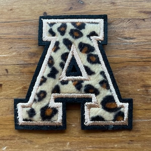 May include: A decorative letter "A" patch with a leopard print design. The letter is outlined in black with a tan border and a cream-colored background. The patch is likely for clothing or accessories.