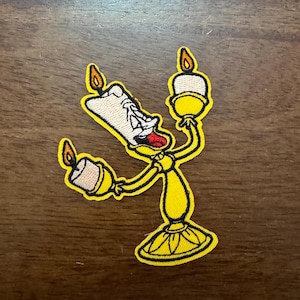 May include: Embroidered patch featuring a yellow anthropomorphic candlestick character with a white candle body, a red tongue, and a smiling expression. The character holds three lit candles with orange flames.