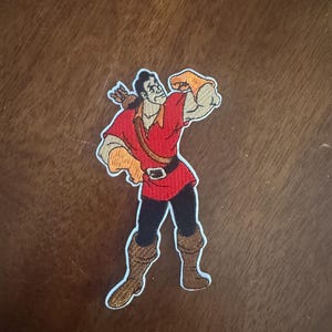 May include: An embroidered patch featuring a cartoon character with dark hair, wearing a red tunic, black pants, and brown boots. The character has a quiver of arrows on their back, wears orange gloves, and is flexing one arm in a pose.