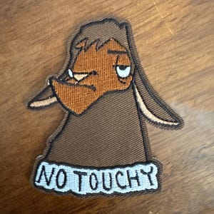 May include: Embroidered patch featuring a cartoon character with a grumpy expression. The character is brown and orange, with the words "NO TOUCHY" in white on a blue background.