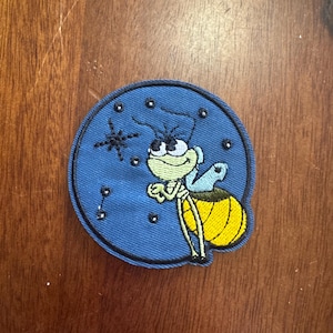 May include: Embroidered patch featuring a cartoon firefly with a yellow abdomen, green body, and blue wings. The patch is circular with a blue background and a star and small dots. The patch is likely for a jacket or backpack.