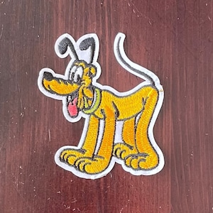 May include: A yellow embroidered patch of Pluto the dog from Disney. Pluto is standing on all fours with his tongue sticking out. The patch is surrounded by a white border.
