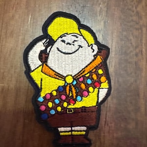May include: Embroidered patch of a cartoon character wearing a yellow shirt, brown pants, and a yellow hat. The character has a white face with a big smile and is wearing a colorful sash.