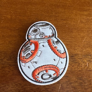 May include: Embroidered patch featuring the BB-8 droid from Star Wars. The patch has a light blue body with orange and black embroidered details. The patch is outlined in black and has a white border.