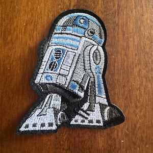 May include: An embroidered fabric patch featuring the iconic R2-D2 droid character. The design is rendered in shades of white, light blue, gray, and black thread, showcasing the robot's cylindrical body, domed head, and mechanical leg details.